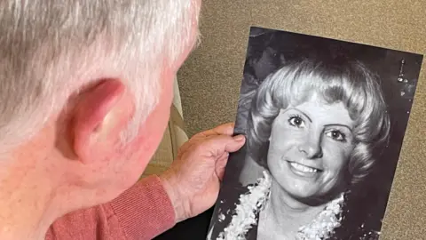 A close shot taken over Jimmy's shoulder as he looks at a beautiful black and white image of his wife, a blonde lady with a broad smile.