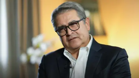 Close up of former Honduran President Juan Orlando Hernandez Alvarado in front of a yellow background. He is wearing a white shirt and black suit 