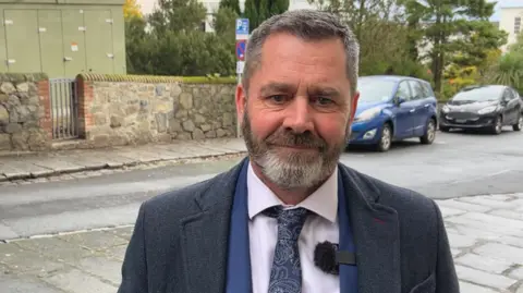 A man with black and grey hair and a black and grey beard. He's wearing a blue suit jacket, with a blue jacket and a white shirt, with a blue patterned tie.
