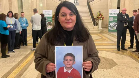 Elena Eke holds a large photo of her son, David. She has long, dark hair and she is looking at the camera with a neutral expression on her face. She is standing in the middle of Stormont's Great Hall.