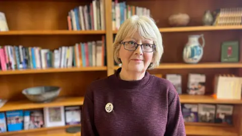 Karen MacKinnon has short, bobbed, blonde hair and is wearing glasses. She is wearing a burgundy high-neck jumper with a gold circular brooch. 