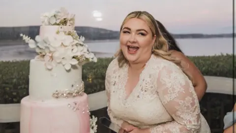 Kayley is wearing a white dress at her wedding and is stood next to a tiered cake, laughing