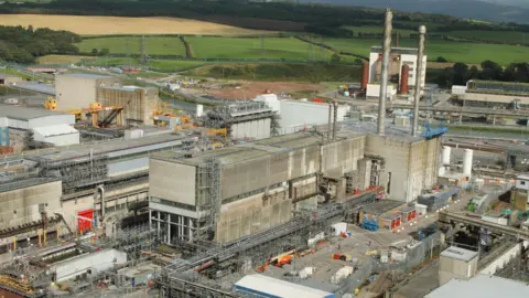 The Magnox Swarf Storage Silo from a distance. It is made up of large grey buildings and two long pipes sticking up into the air. There is lots of machinery nearby. 