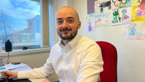 Andrew Turner/BBC Jake Groom is wearing a white shirt and sports a full beard. He has a shaved head, and is sitting at his desk on a red office chair. There is a window behind him which looks out onto Beacon Park in Gorleston and on the wall to the right of the images are pictures drawn by children.