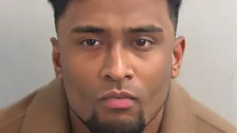 Mug shot of Junead Ahmed, he has a groomed, dark chin beard and dark short hair. He is wearing a caramel wool coat, the top of which can be seen in the image
