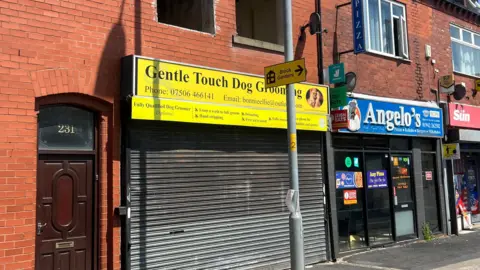 The shuttered front of a dog-grooming salon on a sunny day. On the first floor, there are two empty holes where windows would have been. Other shops as part of the terraced building can be seen in the distance.