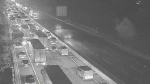 Traffic England/National Highways Traffic queuing on four-lane motorway carriageway - heading away from the camera. Traffic is flowing on the other carriageway.