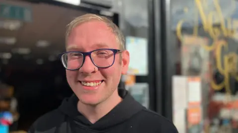 Luke Cook wearing blue glasses and a black hoodie, outside his shop and cafe, Nexus Games