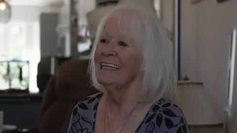An elderly woman with white shoulder-length hair smiles while talking to a person off-camera.