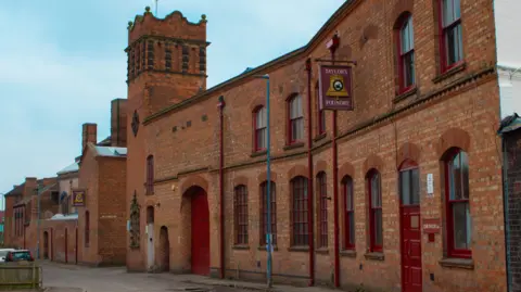 Loughborough Bellfoundry Trust The bell foundry has red doors and red bricks on the outside. A tower is the highest point of the structure. 