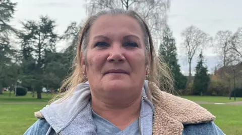 A woman with light brown hair wearing a blue coat standing in a park with trees behind her