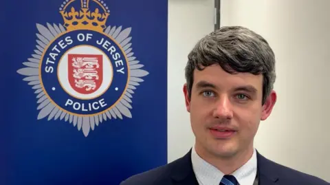 BBC Detective Constable Paul Otter looks to the left of the camera in front of a States of Jersey Police banner. He s wearing a suit.