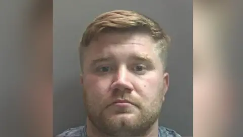 National Crime Agency A custody image of Daniel Pickavance: a man with short light hair and a light beard looking straight ahead. He is wearing a grey, round-neck top.