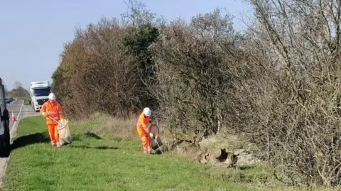 Two people wearing hi-vis clothing litter picking along a major road.