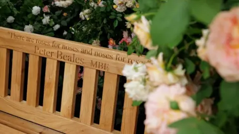 Keith Mindham A wooden bunch covered in roses. Engraved on the bench is a message which is partly covered but the visible writing reads, "There are roses for noses and noses for..." Peter Beales MBE.