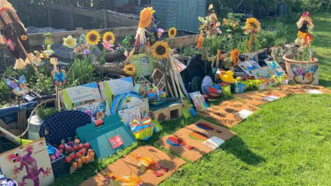 Piles of donated gardening items including mats, kneelers, scarecrows and buckets are placed in front of a school allotment/garden area.