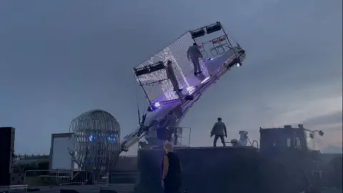 Andrew Turner/BBC On the arm of that crane is a stage, pictured here at a 40% angle with two actors on that stage, attached by safety wire. A third cast member is on the deck of the lorry. The backdrop is the late evening sky. Ultraviolet lights illuminate the stage, which has a background netting of LED lights, with chairs mounted to the upper side panel.