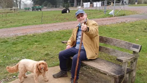 Roy Howard A man holding a walking stick whilst sitting on a bench. A small dog stands next to him and a cow sits in a field behind them. There is a trailer in the background.
