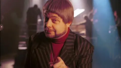 John Thomson as Louis Balfour, presenter of Jazz Club, a sketch featured on The Fast Show - resplendent in a bowl cut, sculpted arty beard, a red polo neck, medallion and a striped jacket.