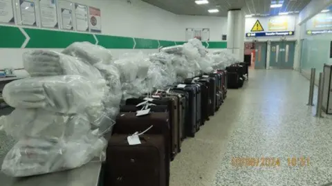 A long row of about 16 brown, blue and black suitcases are lined up with plastic bags dumped on the top of them containing white packets. The bags have been placed in an area of the airport with marble flooring and white walls with green arrows pointing towards a set of two large doors.