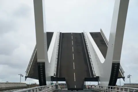 PA Media Drone image of Gull Wing Bridge in Lowestoft. It stretches over green river water and has pedestrian walking at either side of the structure. There are two gery curved accents sticking up into the air from the sides of the bridge, almost in the shape of a boomerang. The river can be seen meeting the sea in the background of the image.