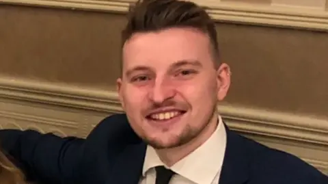 A picture of a man with short, brown, spikey hair, who is wearing a suit and smiling