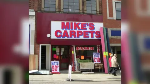 Betty Longbottom/Geograph A shop with a sign saying 'Mike's Carpets'. Outside there are carpet samples and two signs saying 'Mats 50p' and 'Wilton rugs £10'.