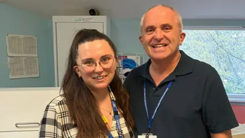 Somerset NHS Foundation Trust Franklin and Reynolds, standing side by side, smiling at the camera, wearing their NHS Trust lanyards and id. Franklin is wearing glasses, a white and black-grey checked blouse and her long brown hair half up and half down.
Reynolds is wearing a black polo shirt, and has short cropped grey hair. They are in front of a window with a trees outside and drawers behind it.