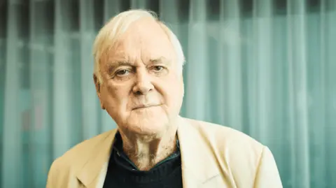 Close-up John Cleese indoors against a green curtain backdrop