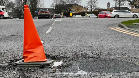 An orange traffic cone placed inside a pothole with a busy mini-roundabout in the background