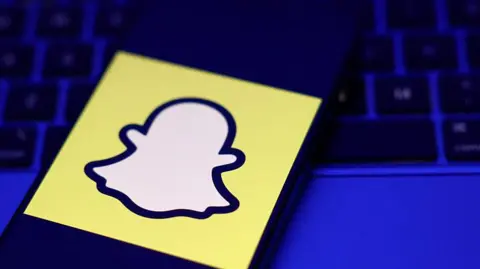 A phone displaying a Snapchat logo on top of a laptop in a darkened room.