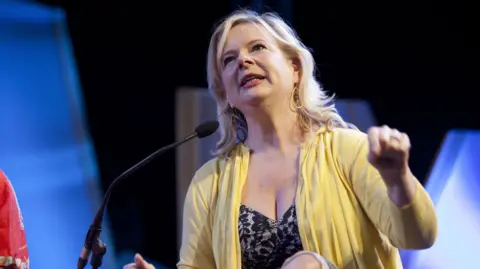 Getty Images Allison Pearson is wearing a black dress and yellow cardi and is speaking into a microphone at the Hay Festival