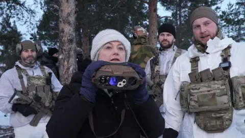 A woman dressed in a dark coat, navy gloves and cream hat stands looking above a pair of binoculars she is holding, She stands surrounded by several soldiers dressed in snow-white uniform, armed with camo belts and guns. The group appear to be in a snowy forest.