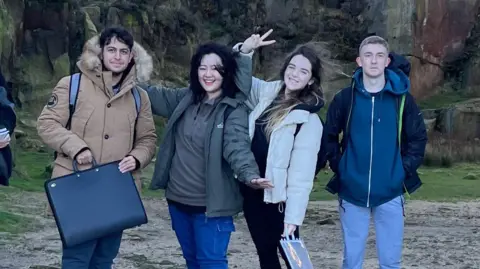 Elina Bodnar Four people - a man with dark hair holding a black briefcase, a woman with dark hair holding her arms out and smiling, a woman with long dark hair making a peace sign with her hand, and a man with light hair with his hands in his pockets - stand alongside each other outdoor with a large rock behind them.
