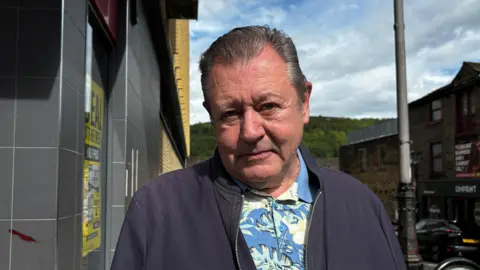 Gemma Dillon/BBC Image shows a man wearing a floral blue shirt and a blue jacket.
In the background is a street scene and hills in the distance. 