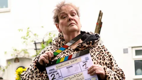 Anne Hegerty is holding a clapper board for Exorcising Barry and looking directly into the camera which is looking up at her. She is wearing a leopard print hooded top. She is standing in a back garden near a white house.