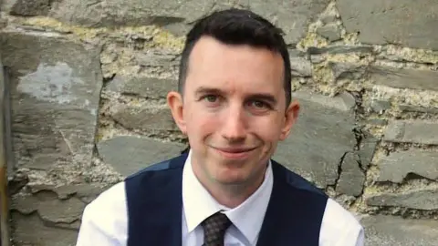 Andrew McCusker Andrew McCusker has short dark brown hair and is wearing a white shirt, dark tie and dark waistcoat. He is smiling in the picture Behind him is a stone wall.