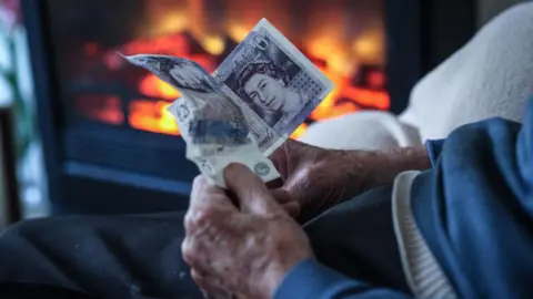 Getty Images An elderly man warms himself in front of a fire. He is holding £20 notes.