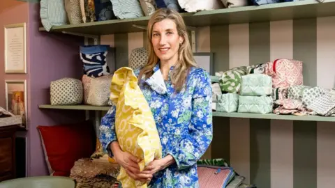 Pinky is standing in front of a shelf full of pillows. She is holding a yellow pillow and wearing a blue, flowery dress. She is smiling at the camera.