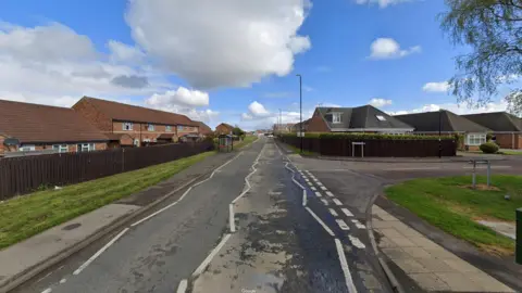 Burdon Road is a single-carriageway road with red-bricked houses behind brown fences bordering it. A metal bus stop is on the left-side pavement. The junction onto Lidcombe Close is on the right.