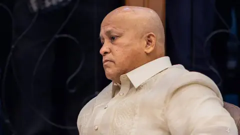 Ronald Dela Rosa, a bald man wearing a cream-coloured button-up shirt, sits and stares at a distance.