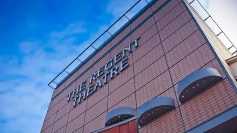 The top half of an orange building with a sign that reads "The Regent Theatre". The sky in the background is bright blue with a few scattered clouds.