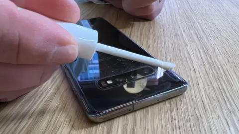 A close-up of a white stick with water on it being applied to a black android phone.