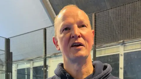 Chris is standing in-front of a black fence. He is wearing a navy hoodie. 