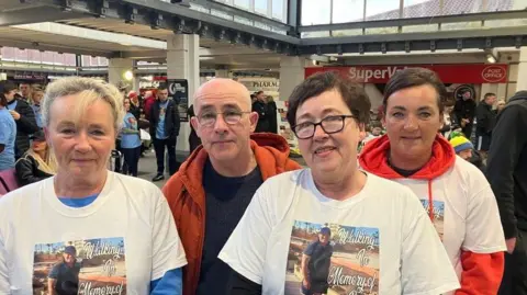 Two women in white t-shirts, along with a man in a brown jacket with grey hair and a black top and a woman with dark hair wearing a white and red top. Maureen McCarthy, front right, has dark hair and glasses.