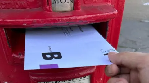 BBC A postal vote being posted by hand into a red letterbox.