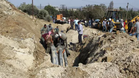 Reuters - Persone ispezionano un'area danneggiata in seguito a quello che i media ufficiali siriani hanno riferito essere un raid israeliano su Masyaf. Sul retro si può vedere un escavatore giallo. E gente che cerca tra le macerie nella sabbia.