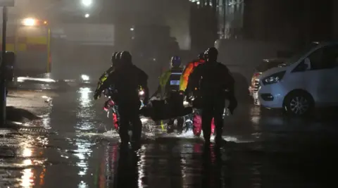 Eddie Mitchell Emergency services at the holiday park in 2024 in the evening carrying a small boat walking through ankle deep water