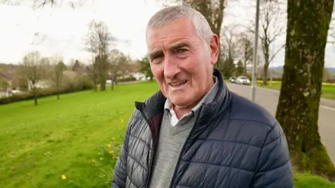 Paul is wearing a blue coat and grey jumper and is standing next to a road overlooking Newport.