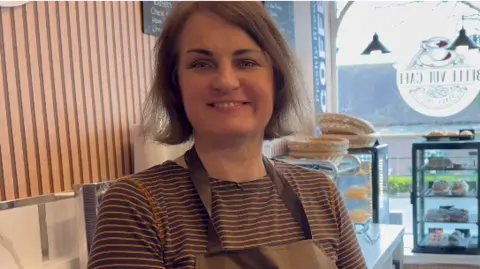 A woman with a light brown bob is wearing a stripped orange and brown top with an apron and is smiling at the camera. She is standing in a cafe